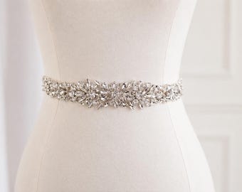 May include: A silver-toned belt featuring an intricate arrangement of clear, faceted crystals and rhinestones. The embellishment is centered and spans horizontally, showcasing a symmetrical floral-inspired pattern with marquise and round stones.