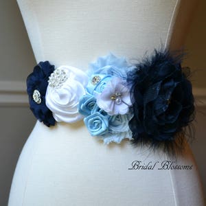 May include: A white, blue, and navy blue satin flower sash with rhinestones and feathers. The sash is tied around a white dress form.