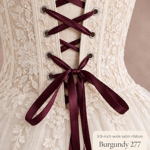 May include: An ivory corset featuring intricate floral lace overlays and a back closure laced with a 3/8-inch wide burgundy satin ribbon. The garment includes metal grommets and a tied bow at the base of the lacing, labeled Burgundy 277.