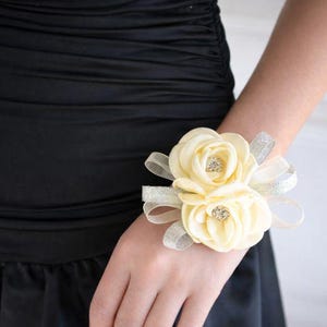 Peut inclure: Un corsage de poignet composé de deux roses en tissu couleur crème avec des centres en strass étincelants, agrémenté de boucles de ruban argenté scintillant, fixé sur un bracelet souple porté sur le poignet contre un vêtement noir.