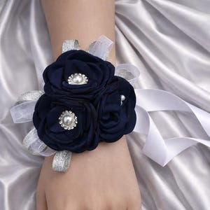 May include: A wrist corsage featuring three dark navy blue fabric flowers with central faux pearl and rhinestone embellishments, accented by silver glittery ribbon loops and long white satin ribbons tied around the wrist.