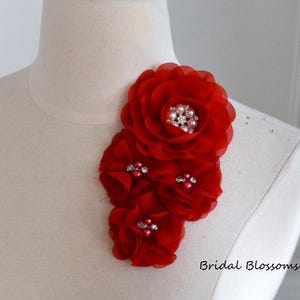 Puede incluir: Adorno floral de tela roja con pedrería y perlas. La flor está hecha de varias capas de tela y está unida a un fondo blanco.