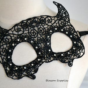 May include: Black lace mask with a devil horn design, adorned with small, sparkling rhinestones. The mask is secured with a black satin ribbon. The mask is displayed on a mannequin, suggesting it's a costume or accessory.