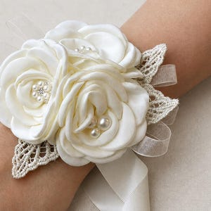 May include: A wrist corsage featuring three cream-colored satin fabric roses, accented with small white pearls and clear crystals in the centers. The floral arrangement is decorated with beige lace leaf cutouts and sheer ribbon loops.