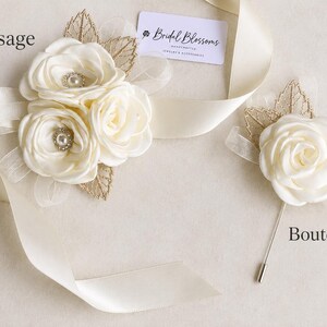 May include: A cream-colored fabric corsage with three roses, pearl centers, and gold leaf accents, alongside a matching single-rose boutonniere with a metal pin. A cream satin ribbon and a tag labeled Bridal Blossoms are included in the display.