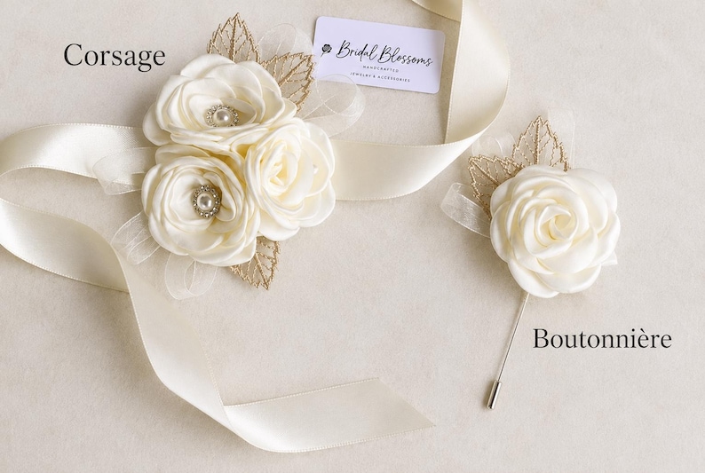 May include: A cream-colored fabric corsage with three roses, pearl centers, and gold leaf accents, alongside a matching single-rose boutonniere with a metal pin. A cream satin ribbon and a tag labeled Bridal Blossoms are included in the display.