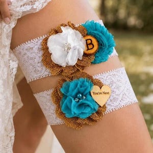 May include: Two white lace garters featuring burlap backing, teal and white fabric flowers, and decorative accents. The top garter includes a wooden circle with the letter B, while the bottom garter features a wooden heart with the text You're Next.