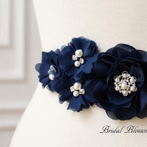 Könnte beinhalten: Ein marineblauer Stoffgürtel mit einer Ansammlung von geschichteten marineblauen Stoffblumen. Das Blumenarrangement ist mit weißen Kunstperlen und klaren Strasssteinen verziert. Der Text Bridal Blossoms ist unten rechts aufgedruckt.