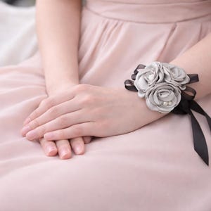 May include: A wrist corsage featuring three light gray fabric roses accented with small white pearls. The floral arrangement is secured to the wrist with a thin black satin ribbon that ties into a bow, creating a contrast between the gray and black.