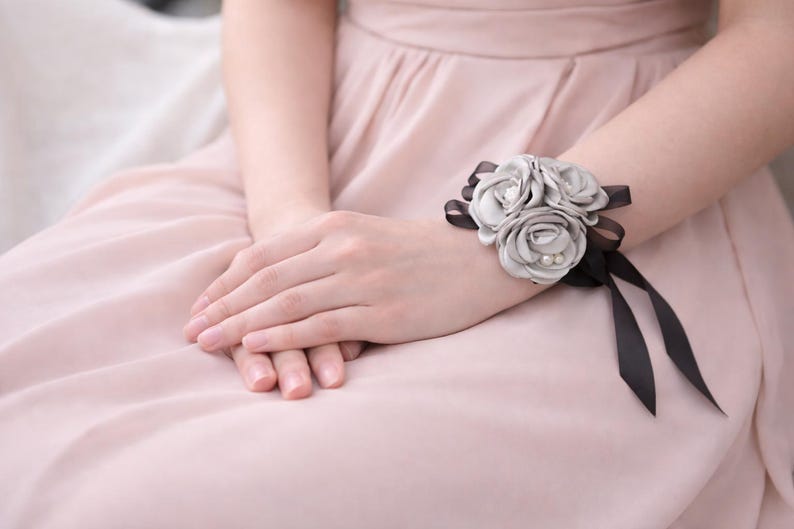 May include: A wrist corsage featuring three light gray fabric roses accented with small white pearls. The floral arrangement is secured to the wrist with a thin black satin ribbon that ties into a bow, creating a contrast between the gray and black.