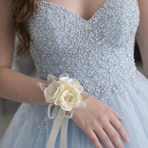 May include: A light blue strapless gown featuring a sweetheart neckline and dense silver beadwork on the bodice, paired with a cream-colored fabric flower corsage worn on the wrist, accented with small clear crystals and thin ribbon streamers.