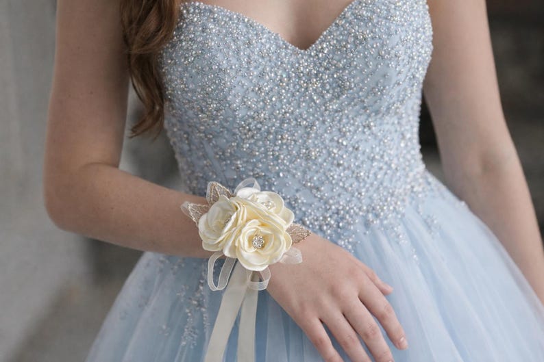 May include: A light blue strapless gown featuring a sweetheart neckline and dense silver beadwork on the bodice, paired with a cream-colored fabric flower corsage worn on the wrist, accented with small clear crystals and thin ribbon streamers.