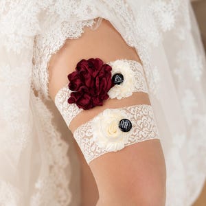 Pode incluir: Duas ligas de renda branca com flores de tecido. A liga superior tem uma flor grande bordô e uma flor pequena creme com um pingente circular preto exibindo um símbolo de triângulo branco. A liga inferior tem uma flor creme e um pingente preto com HP.