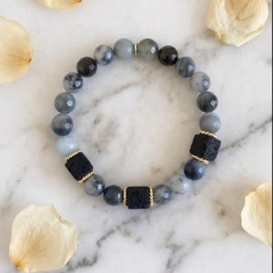 May include: A beaded bracelet featuring a mix of gray, black, and blue-toned round beads. The bracelet includes two black, square lava rock beads with gold-colored accents. The bracelet is displayed on a white marble surface with scattered petals.