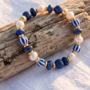 May include: A beaded bracelet featuring a mix of blue, white, and gold accents. The bracelet includes blue lapis lazuli beads, white pearls, and gold-colored spacers. Some beads have a blue and white striped pattern. The bracelet is displayed on a piece of driftwood.