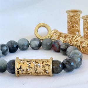 May include: A bracelet composed of faceted grey and dark-toned beads interspersed with a central gold-toned cylindrical bead featuring an intricate floral cutout pattern. Additional gold-toned cylindrical components are visible in the background.