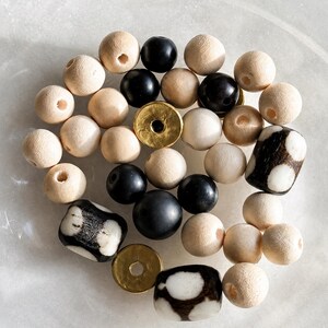 May include: A collection of assorted beads including natural light-colored wooden spheres, smooth black spherical beads, two flat circular gold-toned metal spacers, and three cylindrical beads featuring a black and white spotted pattern.
