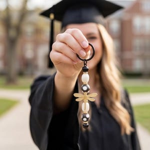 May include: A graduation keychain with a black ring and a beaded design. The keychain features black, white, and gold beads, with a gold dragonfly charm. The background is blurred, showing a person in a graduation gown.