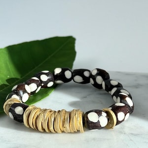 May include: A beaded bracelet featuring dark brown and white polka-dot beads interspersed with gold-colored disc beads. The bracelet is made of natural materials and has a casual, bohemian style.