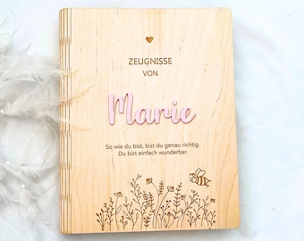 Op de afbeelding: Een lichtgekleurde houten map met een lasergegraveerd ontwerp met een klein hartje, de tekst "ZEUGNISSE VON Marie" en de zin "So wie du bist, bist du genau richtig. Du bist einfach wunderbar." Een bloemenpatroon en een bij zijn onderaan gegraveerd.