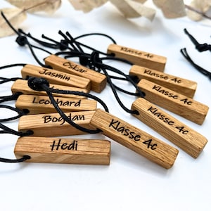 Könnte beinhalten: Holznamensschilder mit schwarzem Band befestigt. Die Schilder sind mit Namen wie "Oskar", "Jasmin", "Leonhard", "Bogdan", "Hedi" und "Klasse 4c" graviert.