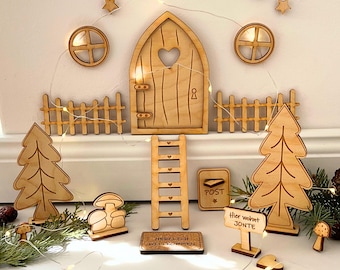 May include: Wooden fairy house decor with a heart-shaped door, a ladder, a mailbox, two trees, a welcome sign, a sign that says "Hier wohnt die Jonté", and two mushrooms. The decor is on a white surface with a string of lights.