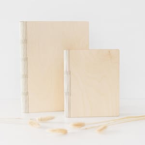 Könnte beinhalten: Zwei hellfarbene Holzkisten in verschiedenen Größen. Die Kisten haben eine natürliche Holzmaserung und sind mit vertikalen schwarzen Linien entlang des Rückens gebunden. Die Kisten stehen auf einer weißen Oberfläche.