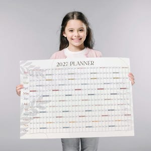 May include: A large wall calendar titled 2027 PLANNER with a grid layout for each month from January to December. The design features a white background with subtle gray leaf illustrations and horizontal rows of colored bars for each month.