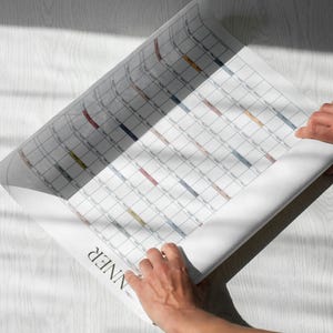 May include: A large white wall planner with a grid layout featuring rows and columns for scheduling. The paper has a matte finish and includes subtle colored accents in various cells. The word PLANNER is printed in black at the bottom.
