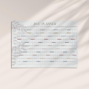 May include: A 2027 wall planner featuring a horizontal grid layout with months listed from January to December on the left side. The planner has a white background with a subtle gray leaf pattern and small colored rectangular accents.