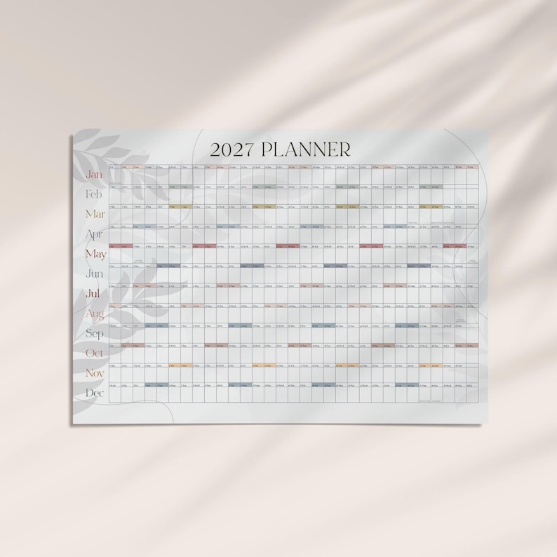 May include: A 2027 wall planner featuring a horizontal grid layout with months listed from January to December on the left side. The planner has a white background with a subtle gray leaf pattern and small colored rectangular accents.
