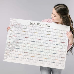 May include: A large wall calendar titled 2027 PLANNER with a white background and a grid layout. The months from January to December are listed on the left side in black text. The design features subtle gray leaf illustrations on the left side.