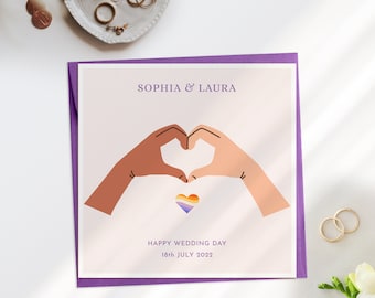 May include: A square wedding card with a purple border features the names "SOPHIA & LAURA" above two hands forming a heart around a rainbow heart. The text "HAPPY WEDDING DAY 18th JULY 2022" is below. Gold rings and jewelry are in the background.