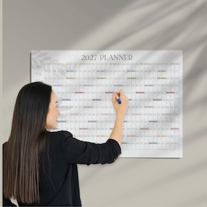 May include: A wall-mounted 2027 planner featuring a grid layout with months listed vertically on the left side and days across the top. The planner has a white background with subtle gray leaf illustrations and colored rectangular accents.
