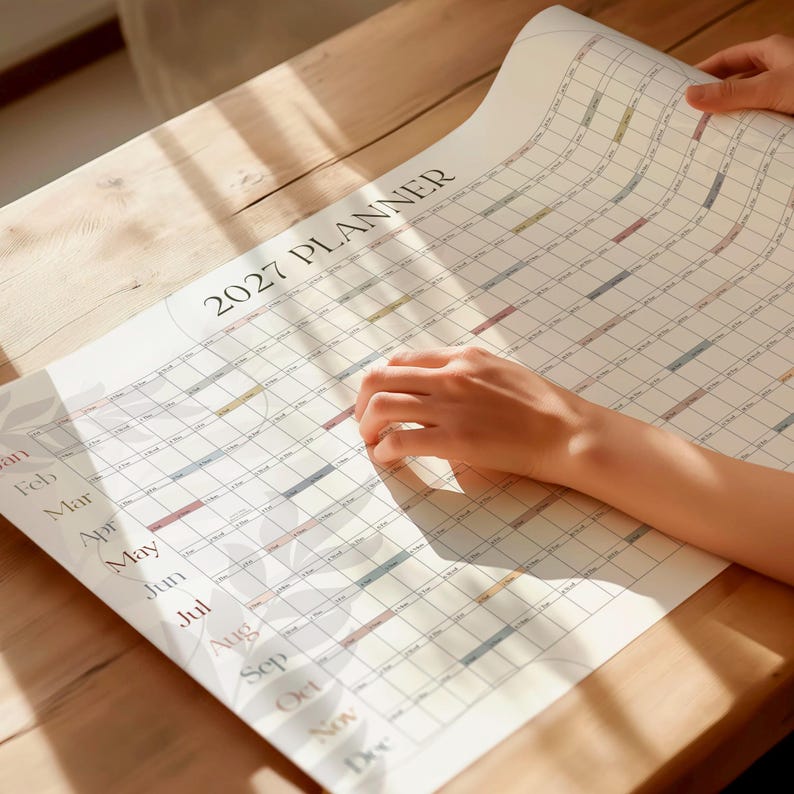 May include: A large wall calendar titled 2027 PLANNER with a grid layout showing months from January to December. The paper is white with black text and subtle pastel-colored accents in the grid cells, featuring a faint leaf pattern design.