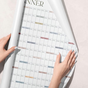 May include: A large white wall planner calendar featuring a grid layout with rows of dates and days of the week. The design includes subtle horizontal bands in muted tones of blue, pink, and tan, with the word PLANNER visible at the top edge.