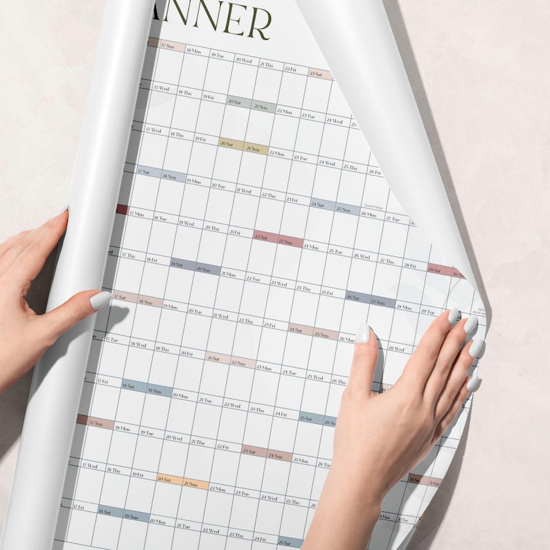 May include: A large white wall planner calendar featuring a grid layout with rows of dates and days of the week. The design includes subtle horizontal bands in muted tones of blue, pink, and tan, with the word PLANNER visible at the top edge.