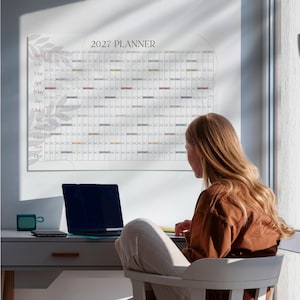May include: A wall-mounted 2027 planner calendar featuring a grid layout with months listed from January to December on the left side. The design includes subtle gray leaf illustrations on the left and right edges, with a white background and black text.