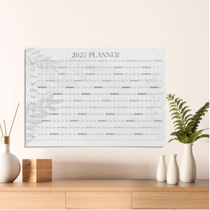 May include: A 2027 wall planner calendar featuring a horizontal grid layout with months listed vertically on the left side. The design includes a subtle light gray leaf pattern in the background, with white grid cells and small colored accents.