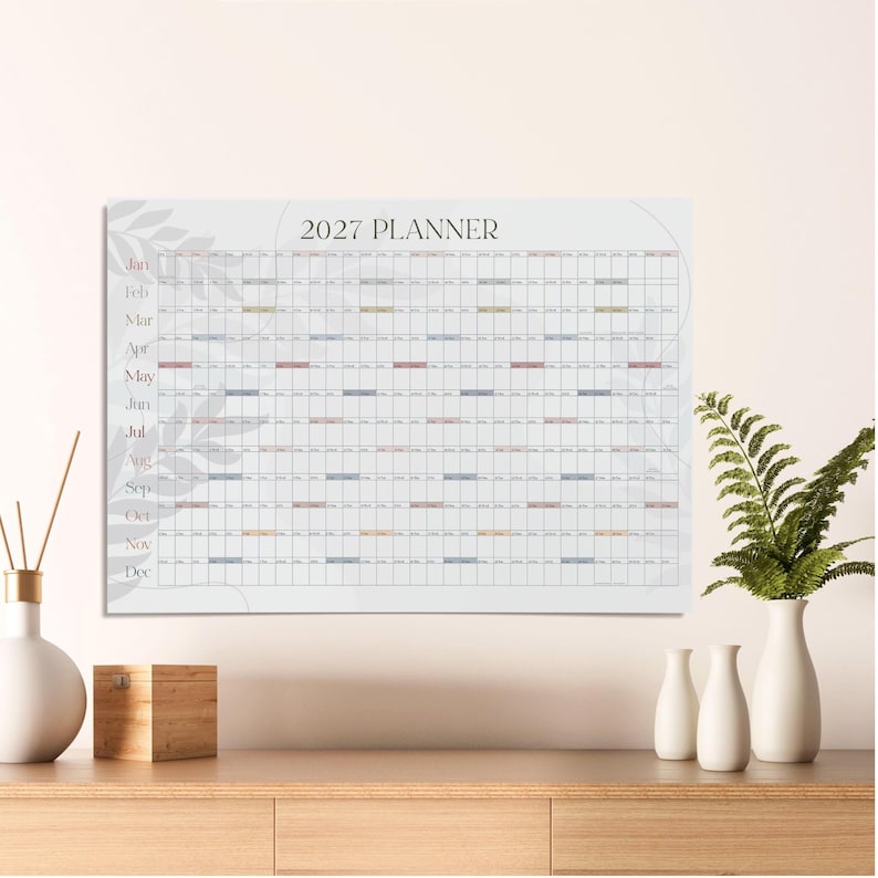 May include: A 2027 wall planner calendar featuring a horizontal grid layout with months listed vertically on the left side. The design includes a subtle light gray leaf pattern in the background, with white grid cells and small colored accents.