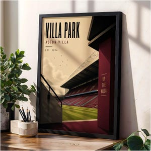 May include: A framed art print featuring a stadium illustration with the text VILLA PARK, ASTON VILLA, EST. 1874, and UP THE VILLA. The graphic depicts a green pitch, red seating, and a dark maroon pillar against a cream-colored sky background.
