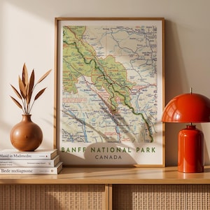 Peut inclure: Une carte encadrée du parc national de Banff, au Canada, avec une disposition topographique détaillée en vert, marron et bleu. La carte est affichée sur un meuble en bois avec une lampe rouge et un vase avec des plantes séchées.