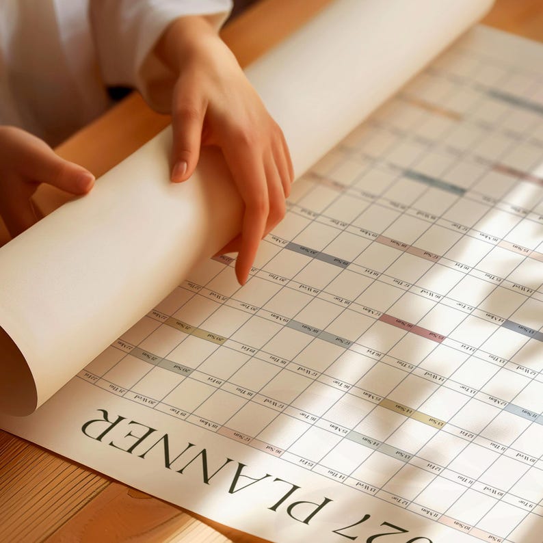 May include: A large, rolled-out paper wall planner featuring a grid layout for the year 2027. The planner has a white background with black text and subtle, muted-colored accents for the days of the week. The word PLANNER is printed in large, bold, black serif font at the bottom of the sheet.