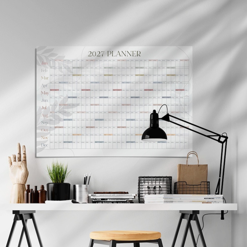 May include: A 2027 wall planner calendar featuring a grid layout with months listed vertically from January to December. The white paper displays a subtle grey leaf pattern in the background. The calendar is mounted on a wall above a desk.