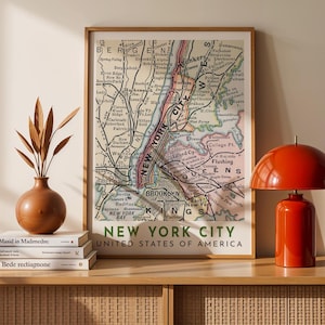 May include: A framed vintage map of New York City, with the text "NEW YORK CITY UNITED STATES OF AMERICA" at the bottom. The map features detailed street names and borough outlines, in shades of beige, pink, and green. The frame is a light brown color.