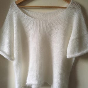 May include: A white short-sleeved sweater with a wide, open neckline and a loose, boxy fit. The garment is crafted from a soft, fuzzy knit material with a visible texture, featuring simple hemmed edges on the sleeves and the bottom of the sweater.