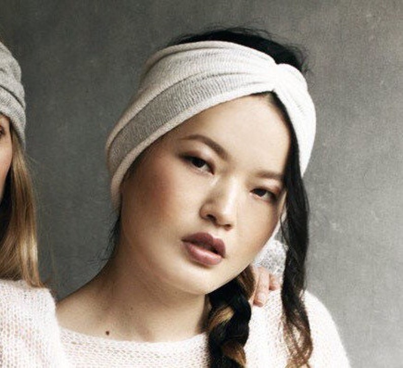 May include: A white and gray knit turban headband with a knotted design. The headband is worn by a person with long black hair.
