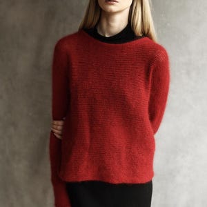May include: A vibrant red long-sleeved sweater with a fuzzy, textured knit finish. The sweater features a simple round neckline and is layered over a solid black base garment. The knit construction is lightweight and semi-sheer in appearance.