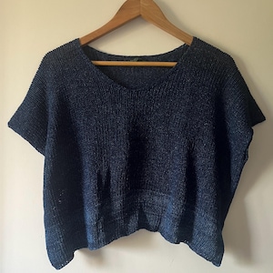 Peut inclure: Un haut en tricot bleu marine avec un col en V et des manches courtes est suspendu à un cintre en bois. Le haut a un aspect texturé et un ourlet légèrement asymétrique. Le tissu semble être de poids moyen.