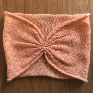 May include: A peach-colored knitted headband with a gathered center detail. The headband is made of a soft, stretchy fabric and is designed to fit comfortably around the head.